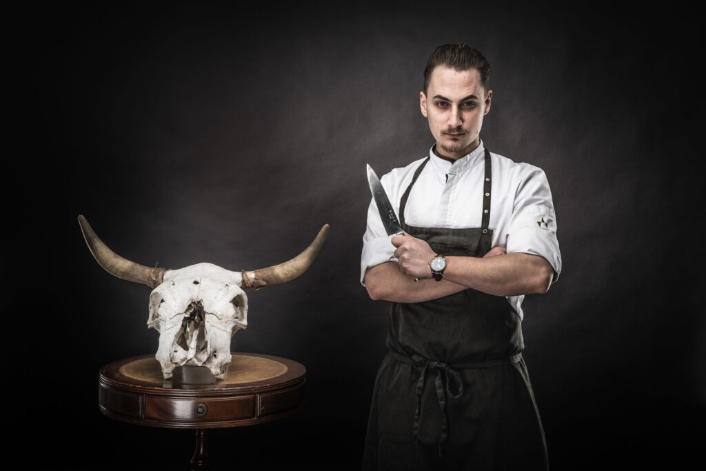 Werbefotografie - Portraitfotografie Florian Wörgötter - Bewerbung für "Junge Wilde 2019" - Schwarzer Hintergrund - Messer in der Hand - ein Stierkopf liegt daneben am Tisch