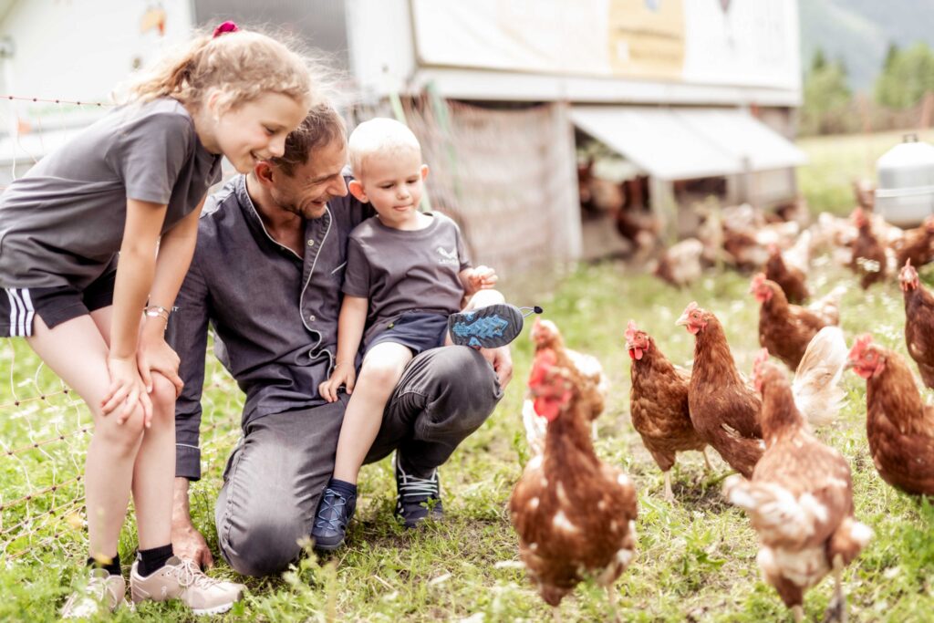 Werbefotografie Hotel Lechzeit - Imageaufnahme des Kochs mit Kindern auf einer Wiese, wo Hühner sind