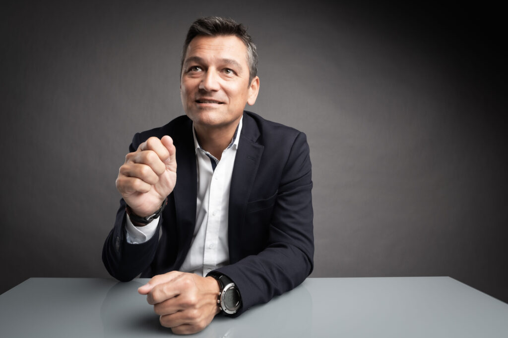 Businessportrait eines Mannes im mittleren Alter. Er sitzt an einem weißen Tisch vor dunkelgrauem Hintergrund. Er gestikuliert mit seiner rechten Hand.