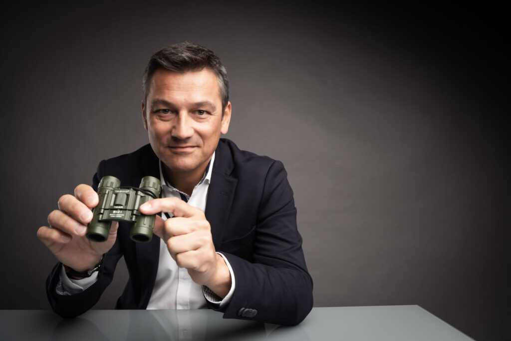 Businessportrait eines Mannes im mittleren Alter. Er sitzt an einem weißen Tisch vor dunkelgrauem Hintergrund. Er hält ein dunkelgrünes Fernglas in seinen Händen.