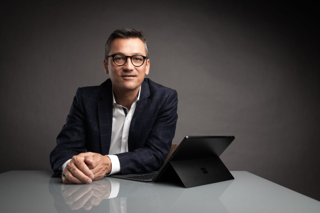 Businessportrait eines Mannes im mittleren Alter. Er sitzt an einem weißen Tisch vor dunkelgrauem Hintergrund. Vor ihm steht ein Laptop und er trägt eine schwarze Brille.