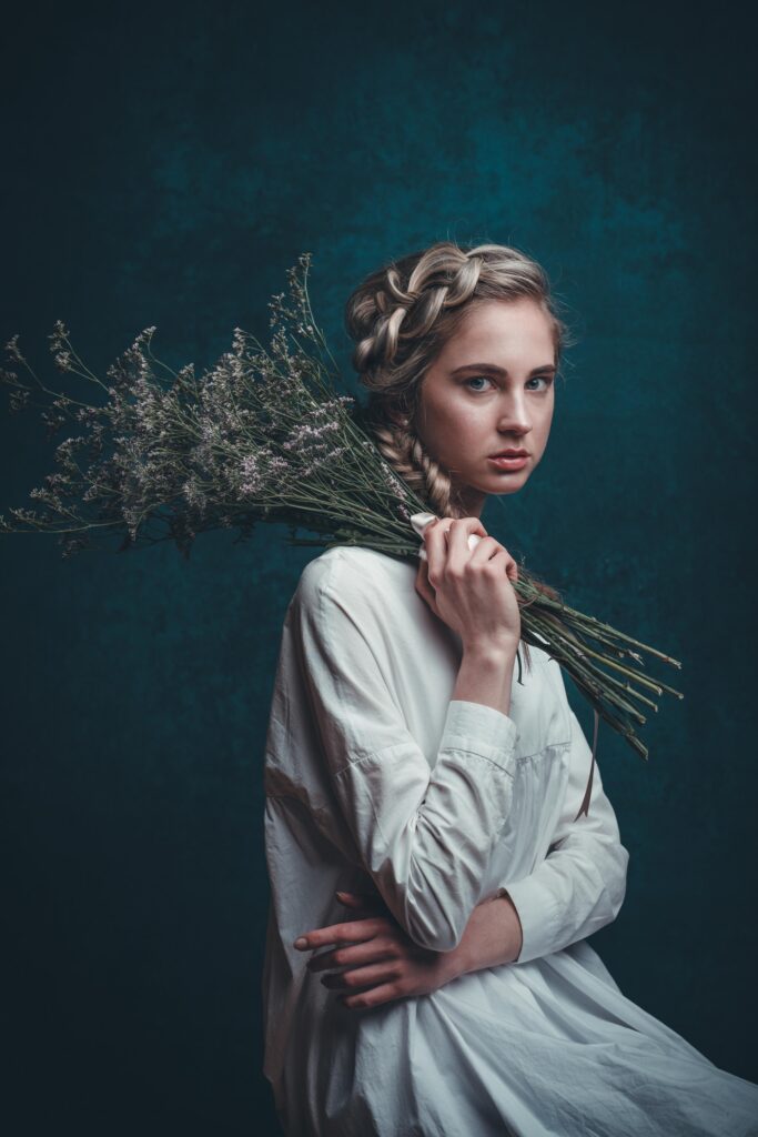 Portraitfoto einer sehr schlanken blonden Frau. Sie hat eine Flechtfrisur, sitzt seitlich und hält einen großen Blumenstrauß über die linke Schulter. Ihr Blick ist sehr ernst. Sie trägt ein weißes Kleid.