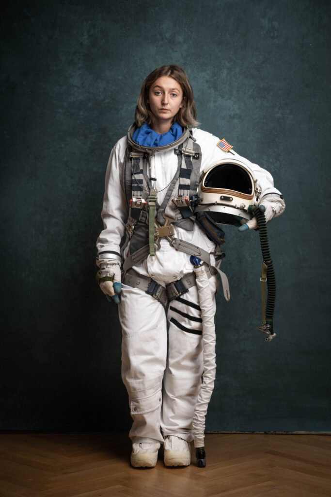 Jana Scherr steht vor einem blauen Hintergrund in einem Astronautenkostüm da. Den Helm hält sie in der Hand