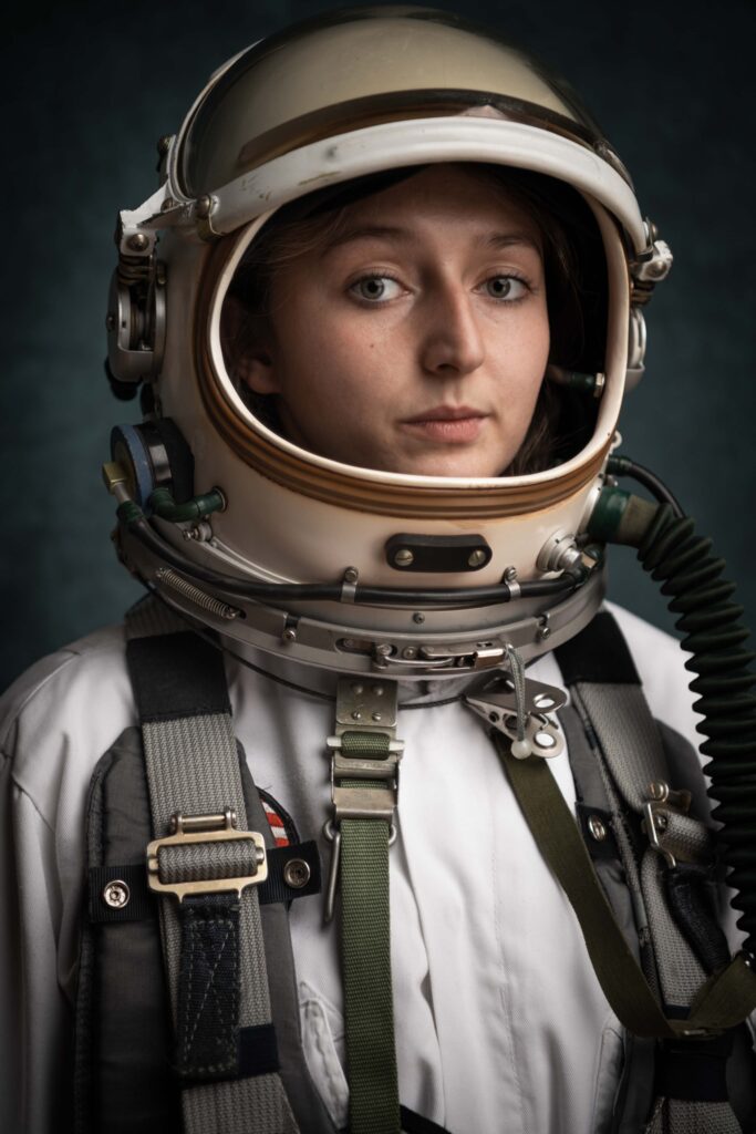 Porträt einer jungen Frau (Jana Scherr) vor blauem Hintergrund. Jana trägt ein Astronautenkostüm.Das Bild ist im Hochformat