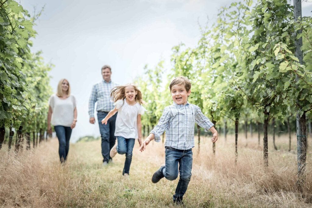 zwei kinder und die Eltern sind in einem Weinberg. Die Kinder laufen glücklich auf die Kamera zu.