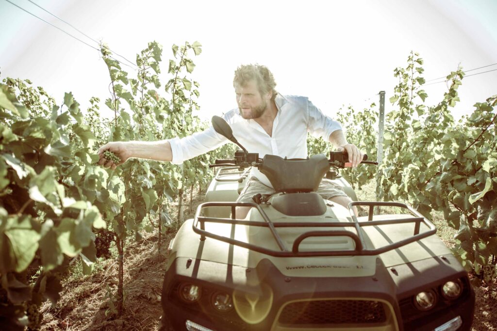Ein Mann (Leo Jahner) fährt mit einem Traktor durch den Weinberg und greift mit seiner Hand in den Rebstock