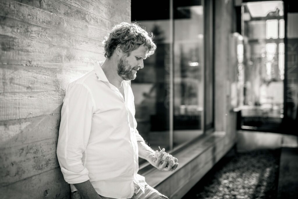 Ein junger Mann lehnt an einer grauen Wand (Wand seines Weinguts)- er hat ein Bein an die wand angewinkelt und wirft lässig weintrauben in die luft. Das Bild ist schwarz/weiß und man sieht den Mann bis zur Hüfte