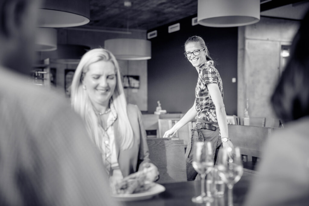 Portrait einer Kellnerin im Buschenschank Jahner - man sieht im Vordergrund verschwommene Menschen an einem Tisch sitzend. Das Bild ist in schwarz/weiß