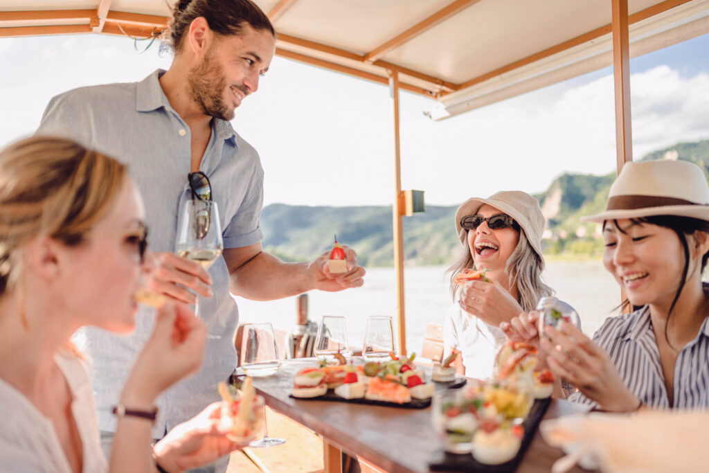 Drei Frauen und ein Mann sitzen auf einem Boot und essen und trinken gemeinsam. Sie haben richtig Spaß.