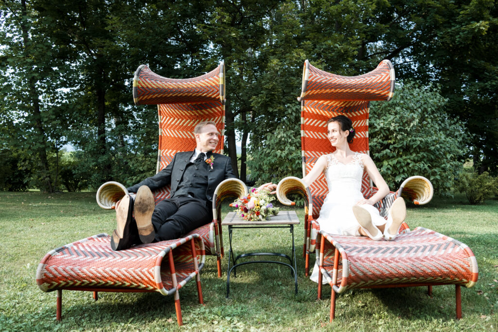 Hochzeitsfotografie- Porträtaufnahme eines Brautpaares, sie liegen in zwei großen roten Ohrensessel in einem Garten