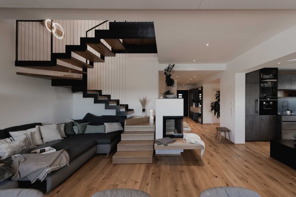 Wohnzimmer mit schwarzer Stiege, brauner Holzboden, graues Sofa und weiße Wände