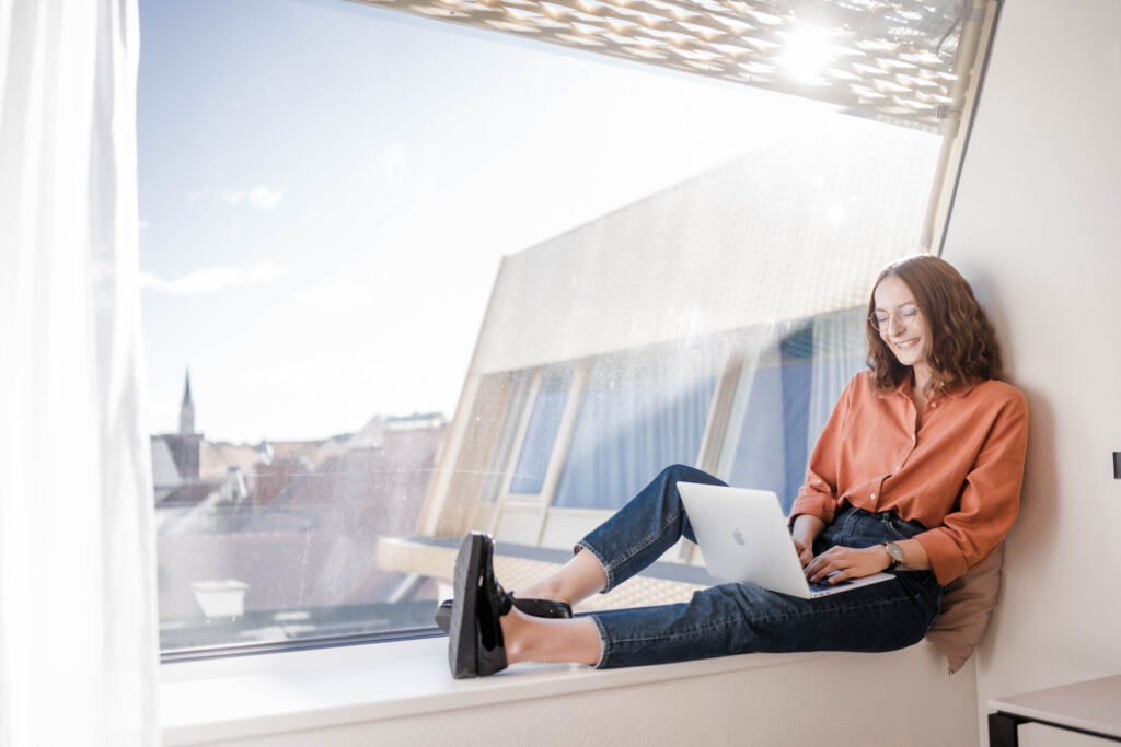 Junge Frau sitzt auf einem Fensterbrett mit einem Laptop am Schoß