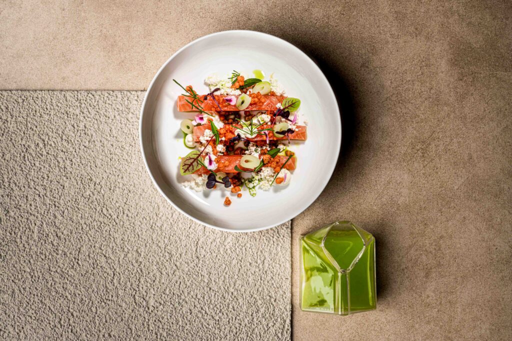 Foodfotografie. Roter Lachs liegt wundefrschön angerichtet und in Streifen auf einem weißen Teller mit hohem Rand. Daneben steht ein Glaskännchen gefüllt mit einem grünen Sud.