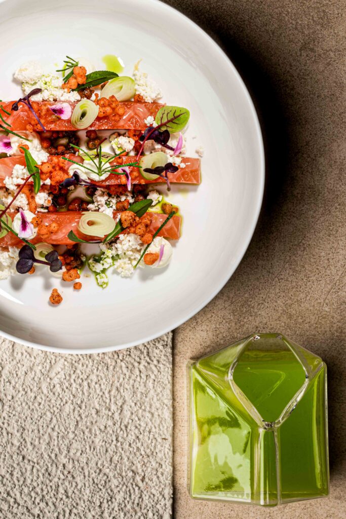 Foodfotografie. Roter Lachs liegt wundefrschön angerichtet und in Streifen auf einem weißen Teller mit hohem Rand. Daneben steht ein Glaskännchen gefüllt mit einem grünen Sud.
