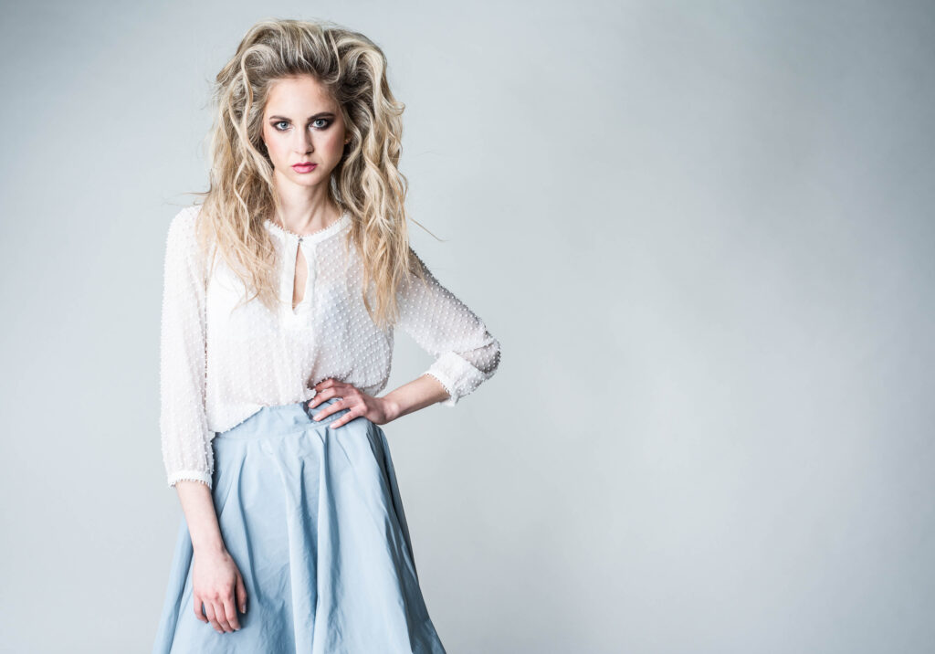 Eine sehr schlanke blonde Frau steht vor einem sehr hellblauen Hintergrund. Sie trägt eine weiße Bluse und ebenso einen hellblauen Rock und hellblaue Highheels. Ihre Frisur ist sehr auffällig- große Locken und toupiert. Man sieht sie am Foto ca. bis zu den Knien. Die rechte Hand hat sie in der Taille eingestützt. die Linke Hand hängt nach unten.