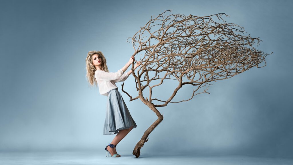 Eine blonde junge Dame steht vor einem hellblauen Hintergrund uns hält einen ausgetrockneten Baum in der Hand. Ihre Beine und der Stamm des Baumes haben dieselbe Biegung.