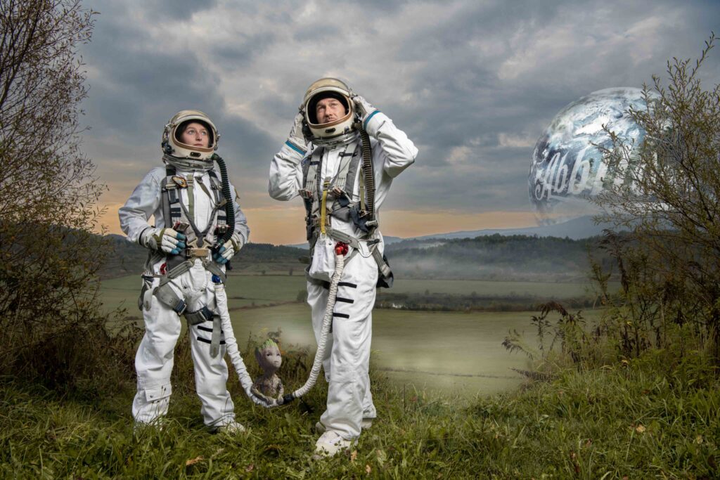 Eine Frau und ein Mann in einem Astronautenanzug stehen in einer grünen Landschaft. Am Himmel sieht man die Erde mit "Die Abbilderei" als Aufschrift.
