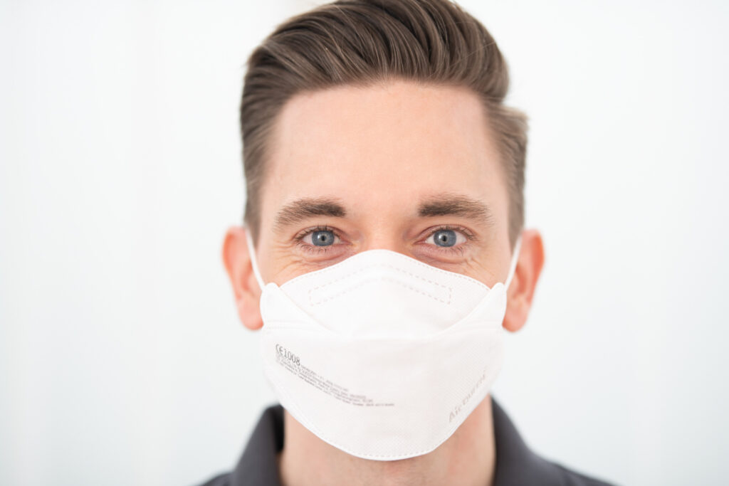 Nahaufnahme eines Gesichts eines jungen Mannes. Er hat hellblaue Augen und trägt einen Mundschutz