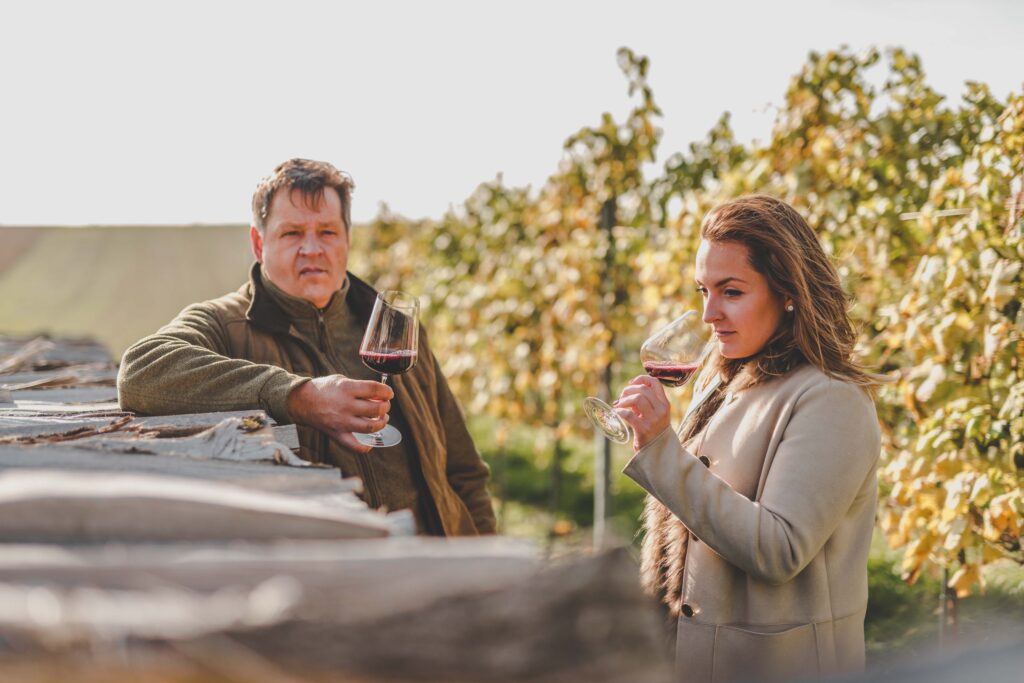 Ein älterer Mann und eine junge Frau stehen im Weinberg und halten ein Glas Wein in der Hand