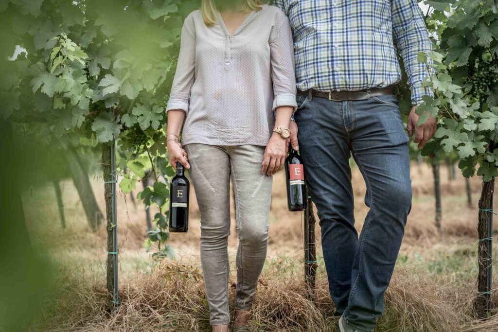 Mann und Frau stehen im Weinberg - man sieht die Körper aber nur bis zu den Schultern. Sie halten eine Flasche Wein in defr Hand