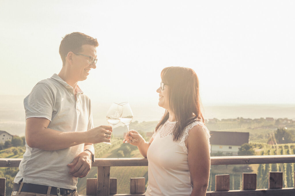 Zwei Menschen (Mann und Frau) stehen an einem Holzbalkon und stoßen mit einem Glas Wein an