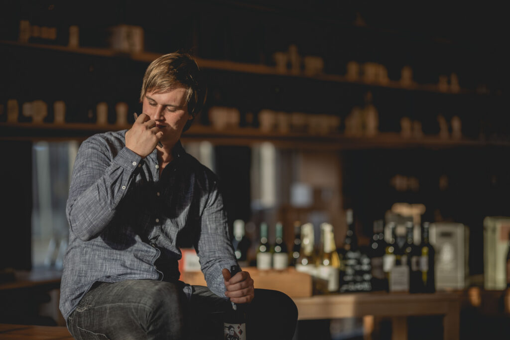 Ein junger Mann (Winzer) sitzt auf einem Tisch in einer Vinothek und riecht an einem Weinkorken.