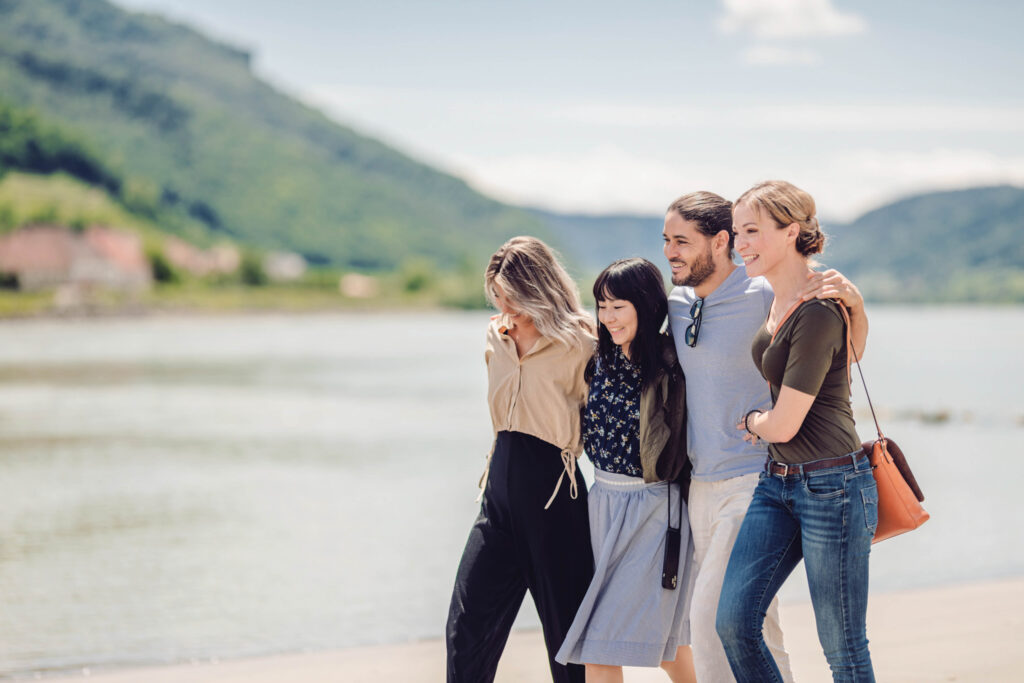 Man sieht 4 Menschen- 3 Frauen einen Mann- an einem breiten Flussufer miteinander Spaß haben