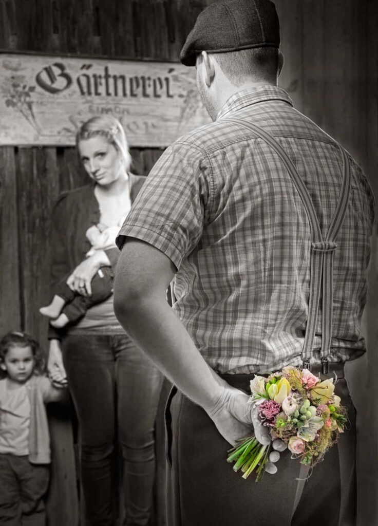 Ein Mann steht mit dem Rücken zur Kamera und hält hinter seinem Rücken einen Blumenstraß. Vor ihm stehen seine Frau undf seine Tochter.