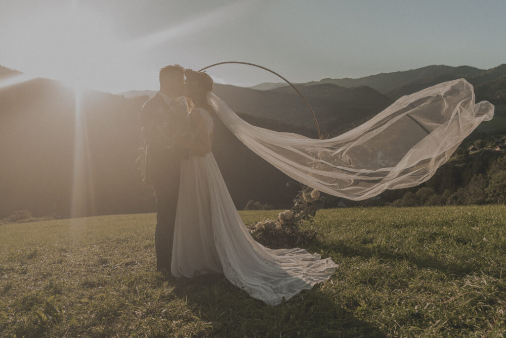 Hochzeitsfotografie- Portraitaufnahme eines Brautpaares von einem mit Blumen geschmückten RIng- das Paar küsst sich und der Schleier weht im Wind